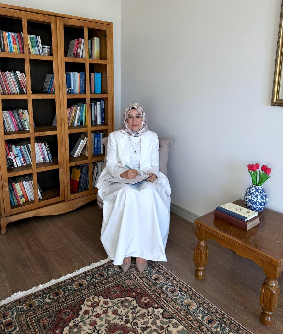 Huriye Çevik, kitaplığın önünde koltukta oturmuş not alıyor; yanında lale dolu vazo